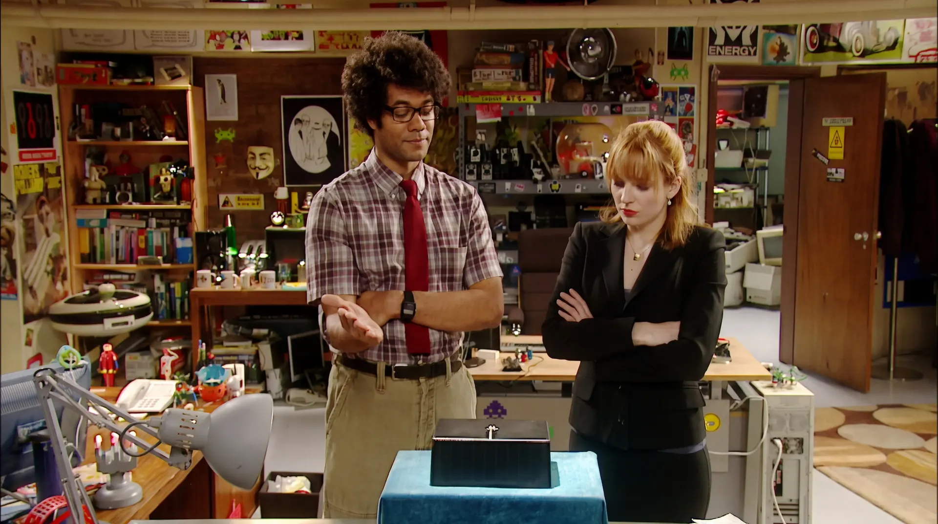 Moss presenting a small black box on a blue cushion while Jen looks on.